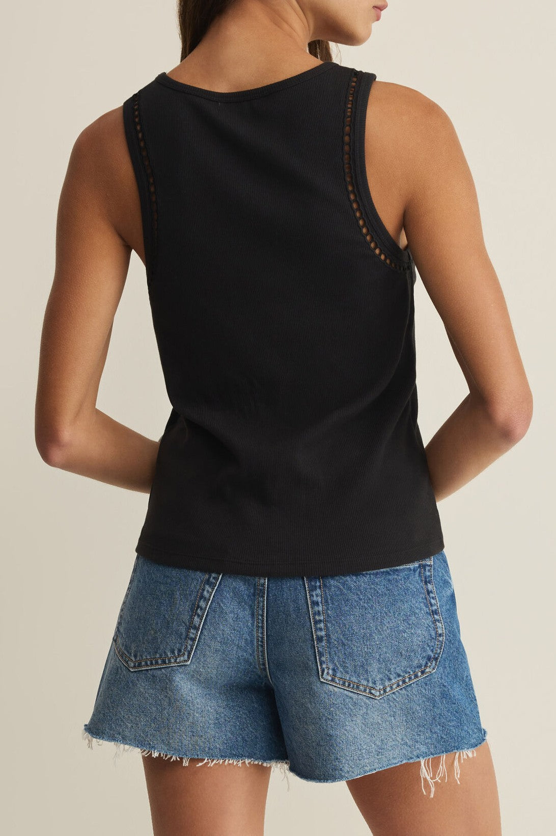 Someone stands with their back to the camera against a light background, wearing the Z Supply Boa Rib Tank in black—a sleeveless crew neck top—paired with blue denim shorts featuring frayed hems.