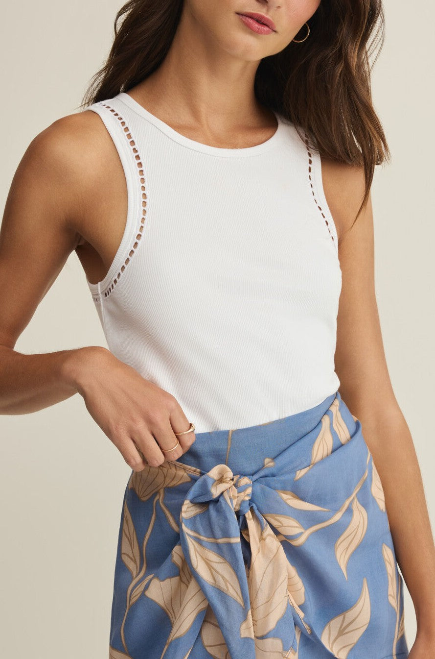 A woman with wavy brown hair wears the Z Supply Boa Rib Tank in white, featuring eyelet details at the shoulders, paired with a blue skirt with beige leaf print tied at the waist. She poses with one hand on her waist.