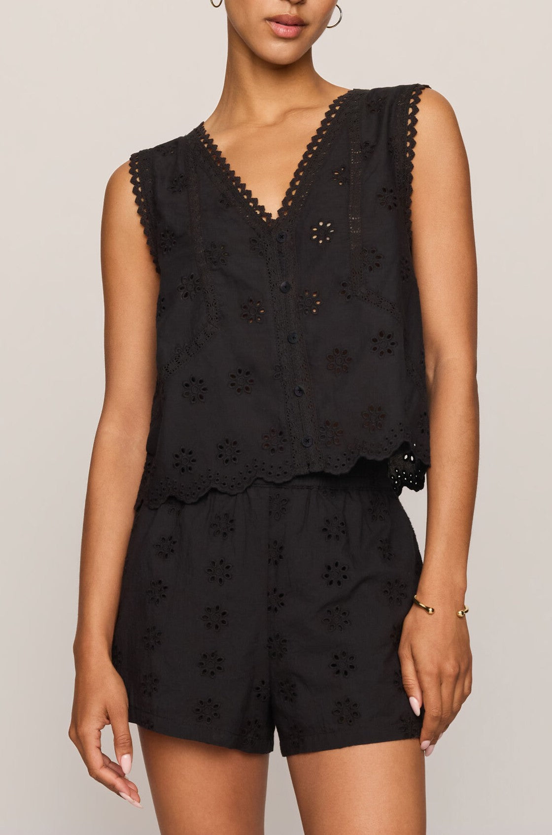 A woman wears the Z Supply Darcy Top in BLACK—a sleeveless, 100% cotton eyelet style with scalloped edges—paired with matching high-waisted shorts. She stands against a neutral background, accessorized with hoop earrings and a bracelet.