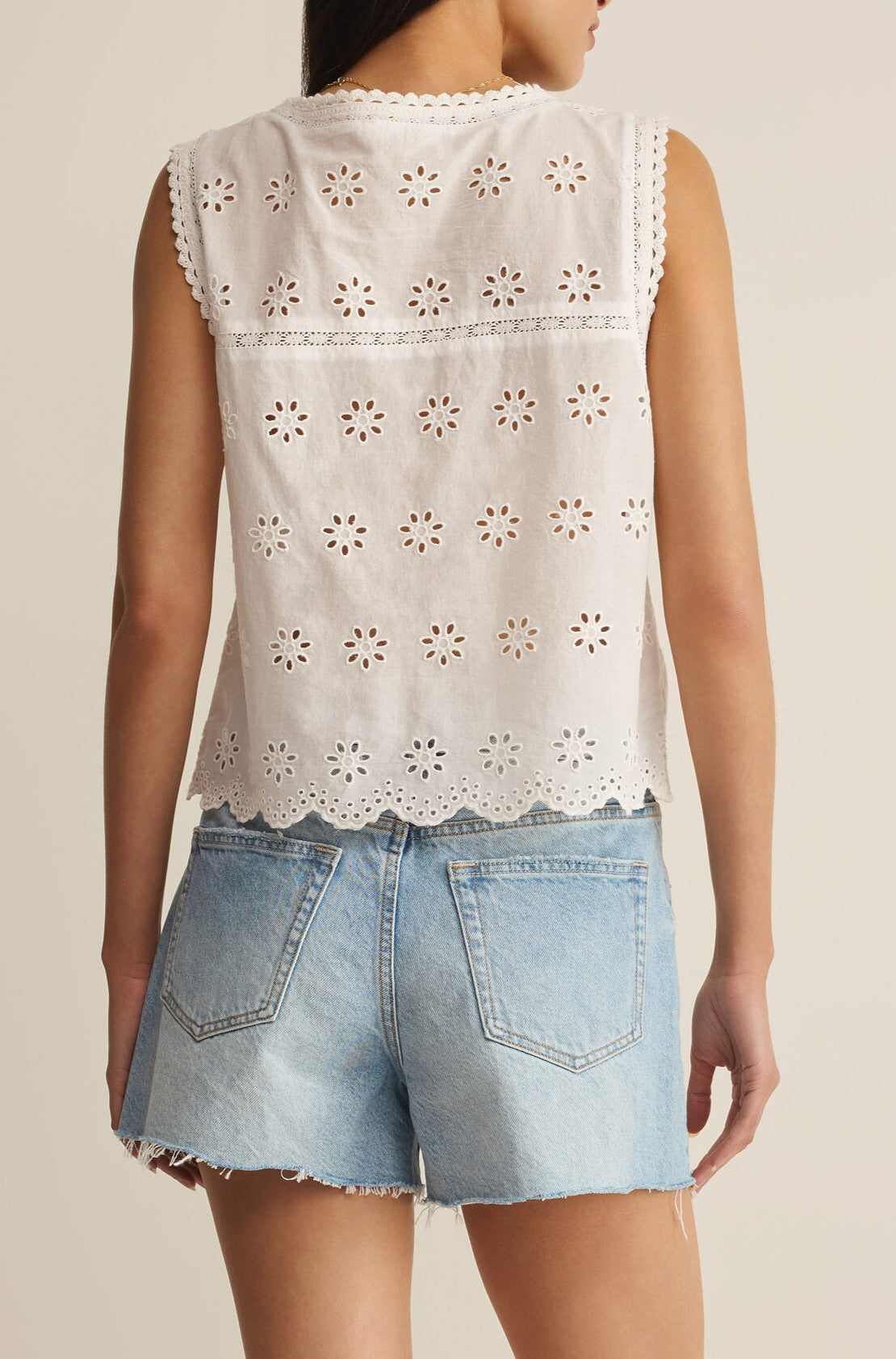 A person is shown from the back wearing the Z Supply Darcy Top—a sleeveless, V-neck white eyelet lace top made from 100% cotton—paired with light blue frayed denim shorts, set against a neutral background.