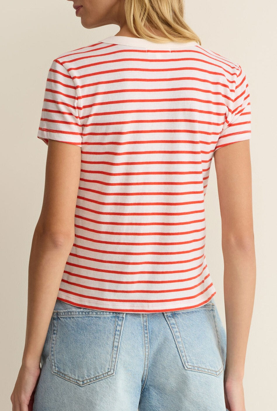 A person with long blonde hair wears the Z Supply Junie Striped Tee—white with red stripes in premium brushed slub jersey cotton—and light blue high-waisted jeans, shown from behind against a neutral background.