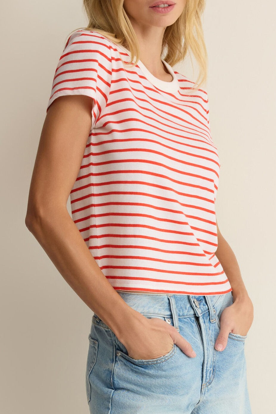 A woman stands with her hands in her pockets, wearing the Z Supply Junie Striped Tee in Red Multi and light blue jeans, set against a neutral background.