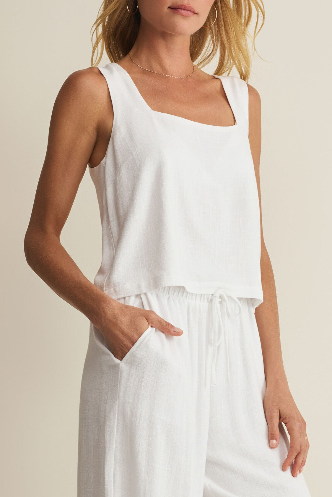 A woman wears the Z Supply Charli Top, a regular fit square neck sleeveless white top, with matching drawstring pants, one hand in her pocket, standing against a neutral background.