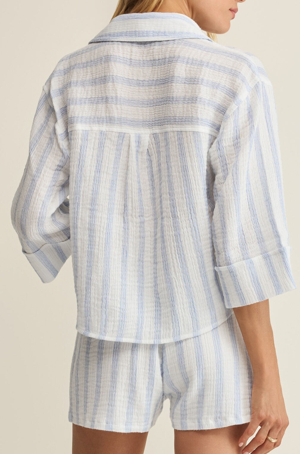 A person stands facing away, wearing the Helena Striped Shirt by Z Supply—a light blue and white stripe blouse made from lightweight, textured fabric.