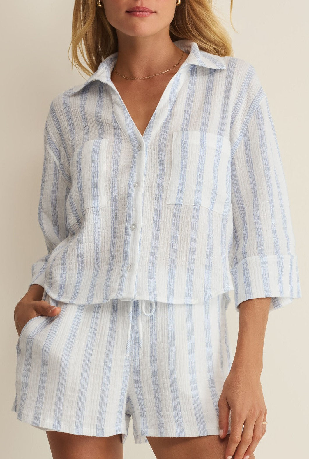 Model wears the Helena Striped Shirt by Z Supply in white—a light blue and white striped, long-sleeve, slightly cropped blouse with a relaxed fit, paired with high-waisted shorts. One hand is in her pocket for a casual look.