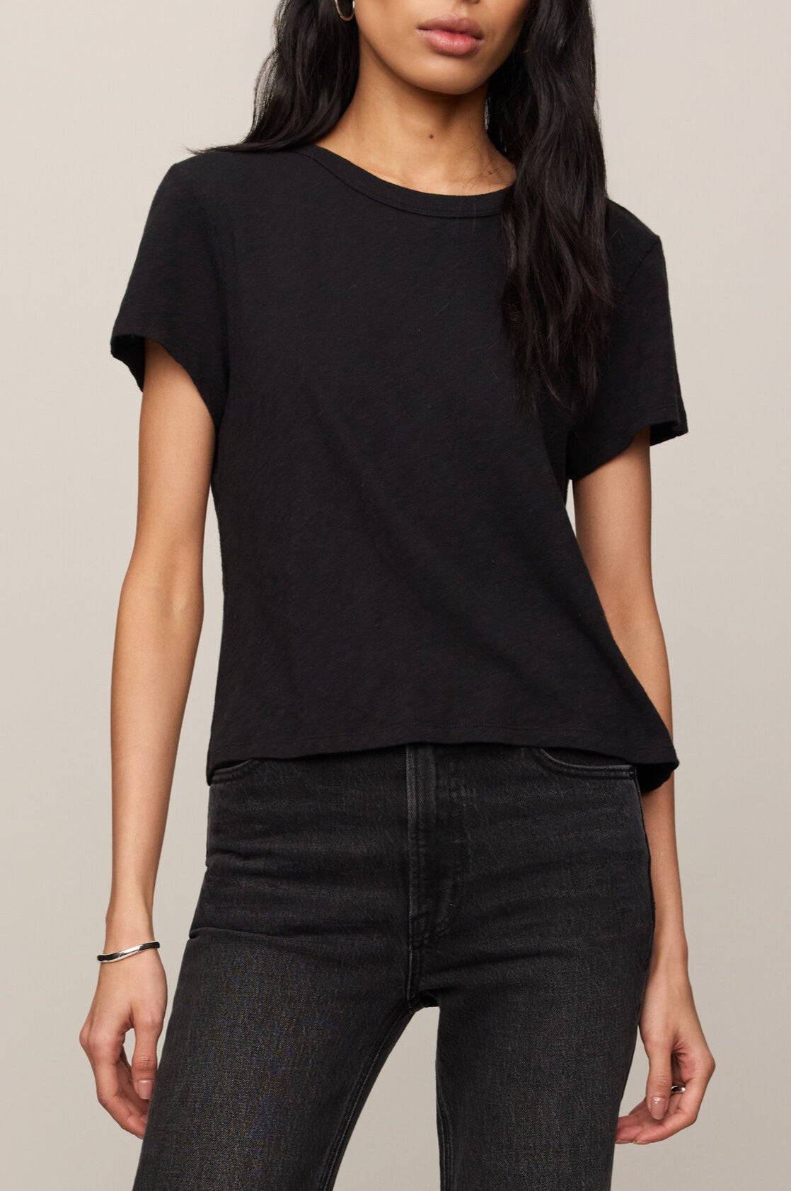 A woman with long dark hair wears the Junie Slib Tee Shirt by Z Supply in BLACK—a regular fit crew neck t-shirt made from 100% cotton—paired with dark jeans, posing against a neutral background with one arm relaxed by her side.