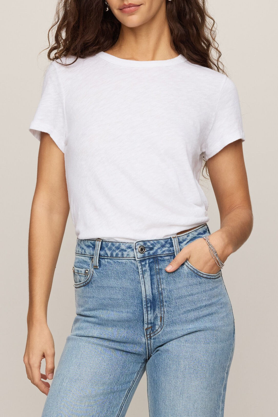 A woman wears the Z Supply Junie Slib Tee Shirt in WHITE, a plain 100% cotton crew neck style, tucked into high-waisted light blue jeans. Her long wavy hair falls over her shoulder, one hand rests in her pocket, and she accessorizes with silver bracelets.