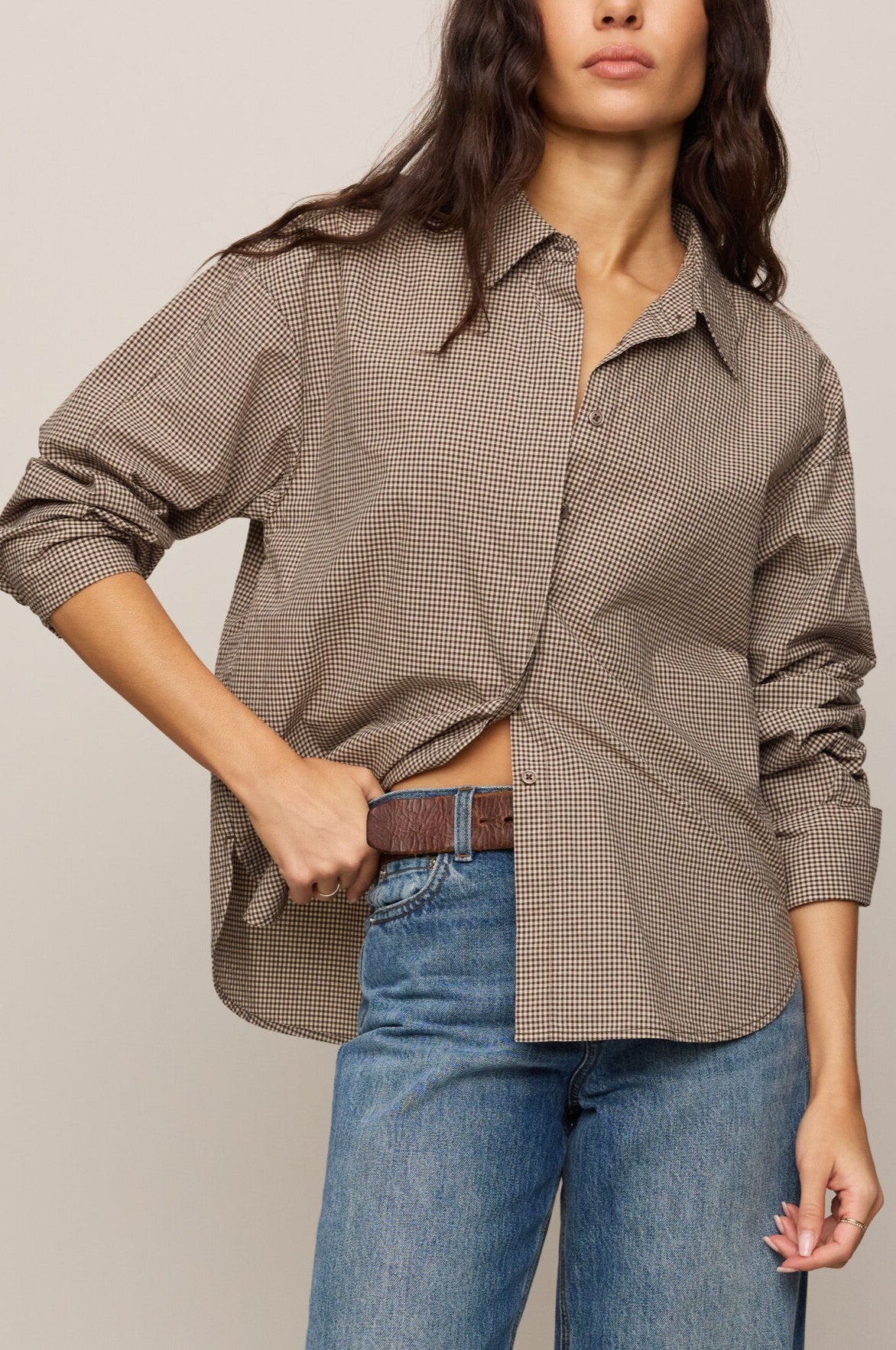 A person wearing the Z Supply On Duty Gingham Button Up in Dark Chocolate stands with one hand in their pocket against a neutral background. Their face is partially visible, and they pair the shirt with blue jeans and a brown belt.