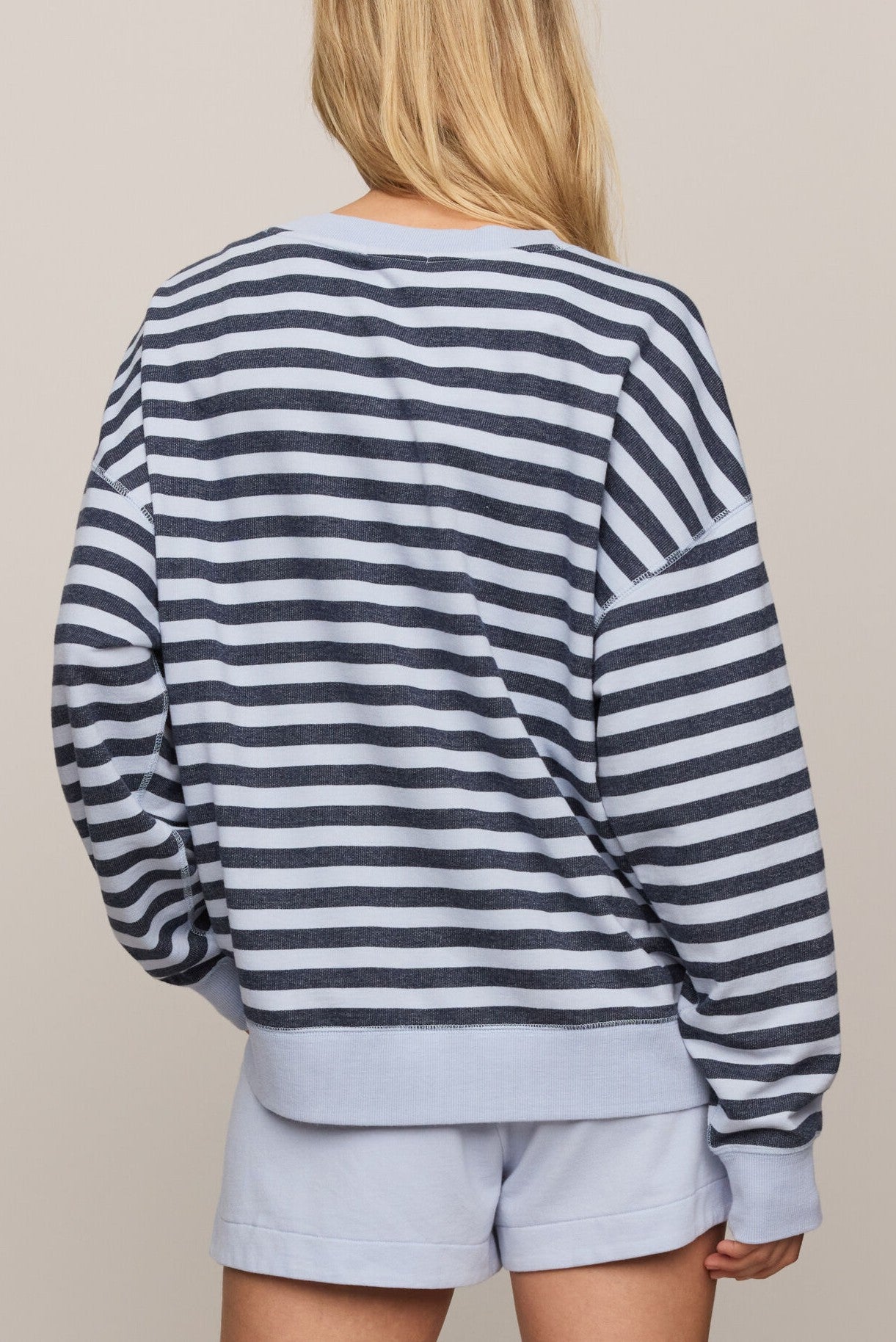 A person with long blonde hair, seen from behind, wears the Z Supply "The Everyday Striped Crew In Eclipse," a navy and white striped oversized long-sleeve crew neck top, paired with light blue shorts against a neutral background.