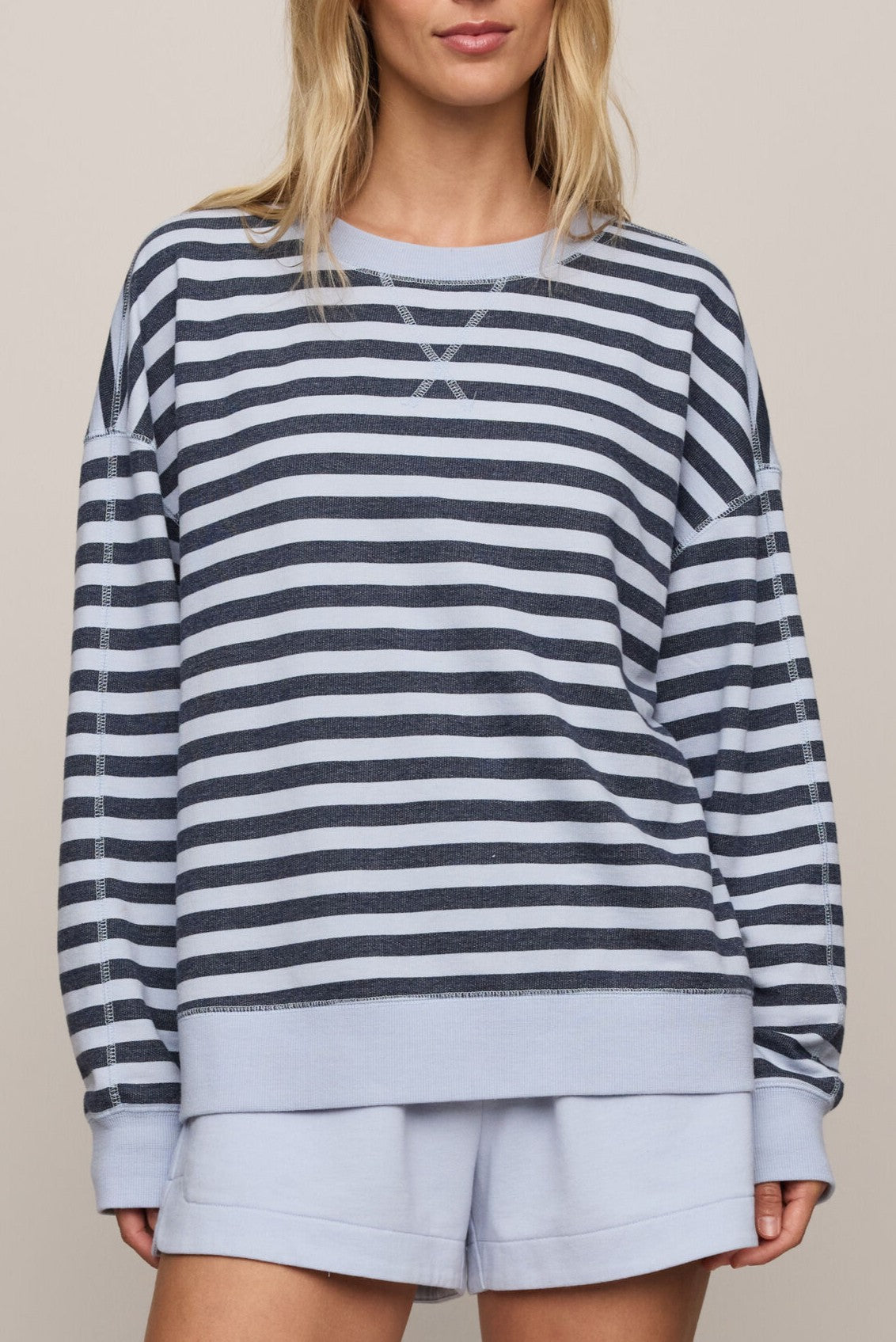A person wearing Z Supply's The Everyday Striped Crew in Eclipse—a long-sleeved, blue and white striped sweatshirt with an oversized fit and crew neck—paired with light blue shorts, stands against a plain beige background. The person's face is partially out of frame.