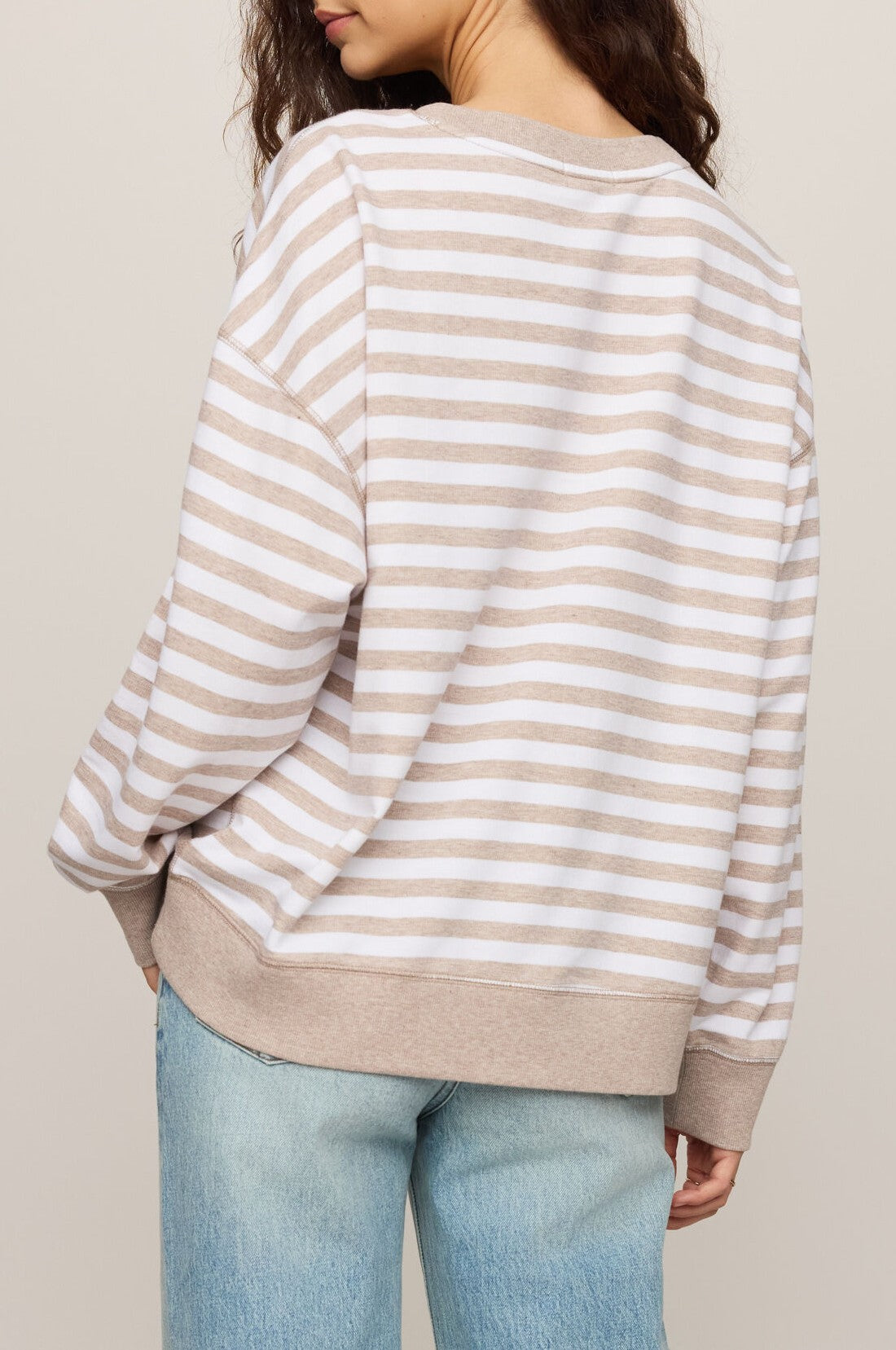 A person with wavy brown hair is shown from the back against a plain background, wearing Z Supply’s The Everyday Striped Crew in Heather Ash—a beige and white striped, oversized fit long sleeve crewneck sweatshirt—paired with light blue jeans.