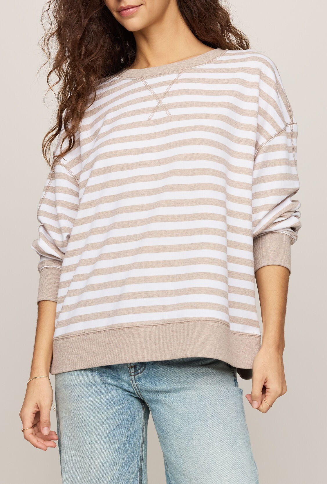 A person wearing The Everyday Striped Crew In Heather Ash by Z Supply—a loose, oversized-fit sweater in HEATHER ASH with beige and white stripes, long sleeves, and a classic crew neck—paired with light blue jeans and standing against a neutral background.
