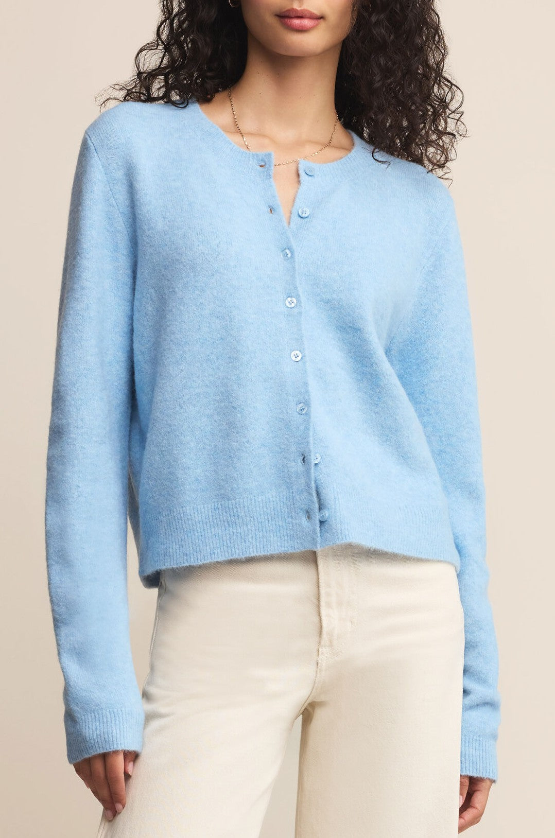 A woman stands against a neutral background wearing the Medina Cardigan in Serene Blue by Z Supply over cream-colored pants. Her curly hair falls around her shoulders, partially obscuring her face.
