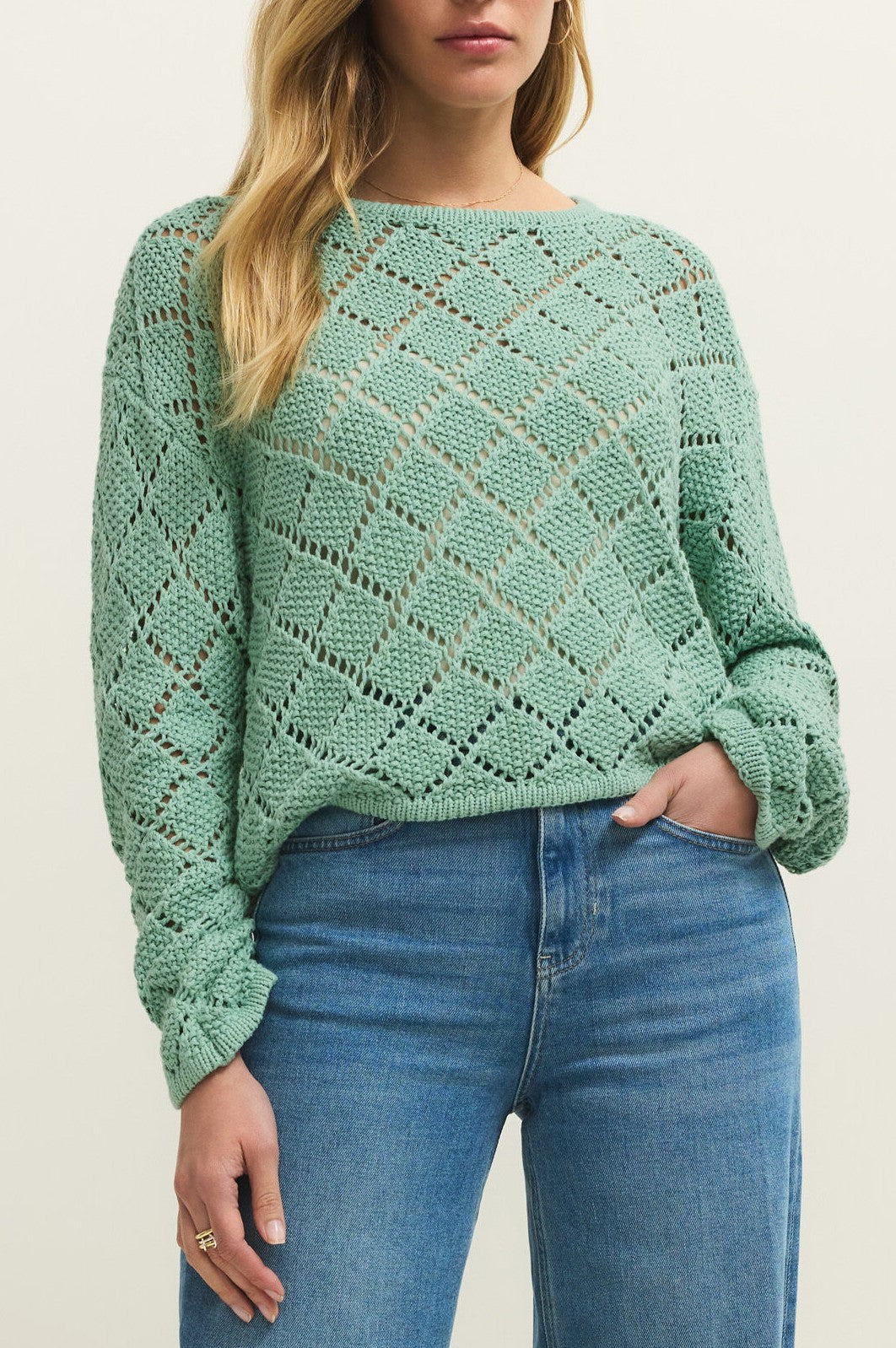 A woman wears the Z Supply Hullen Crochet Sweater in light green with long sleeves and a diamond pattern, paired with light blue high-waisted jeans. She has long, wavy blonde hair and stands against a plain background.