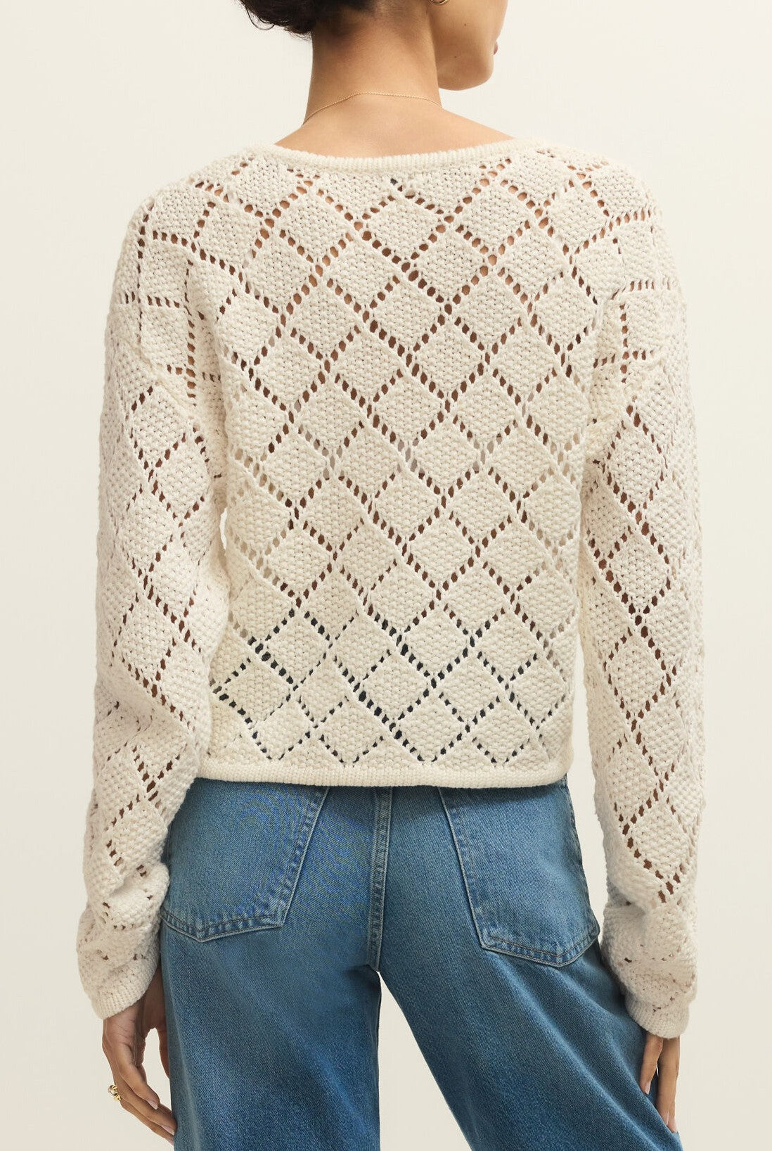 A person is shown from behind wearing the Z Supply Hullen Crochet Sweater, a cream-colored, long sleeve cotton top with a loose fit, detailed diamond lattice pattern, and paired with blue jeans.