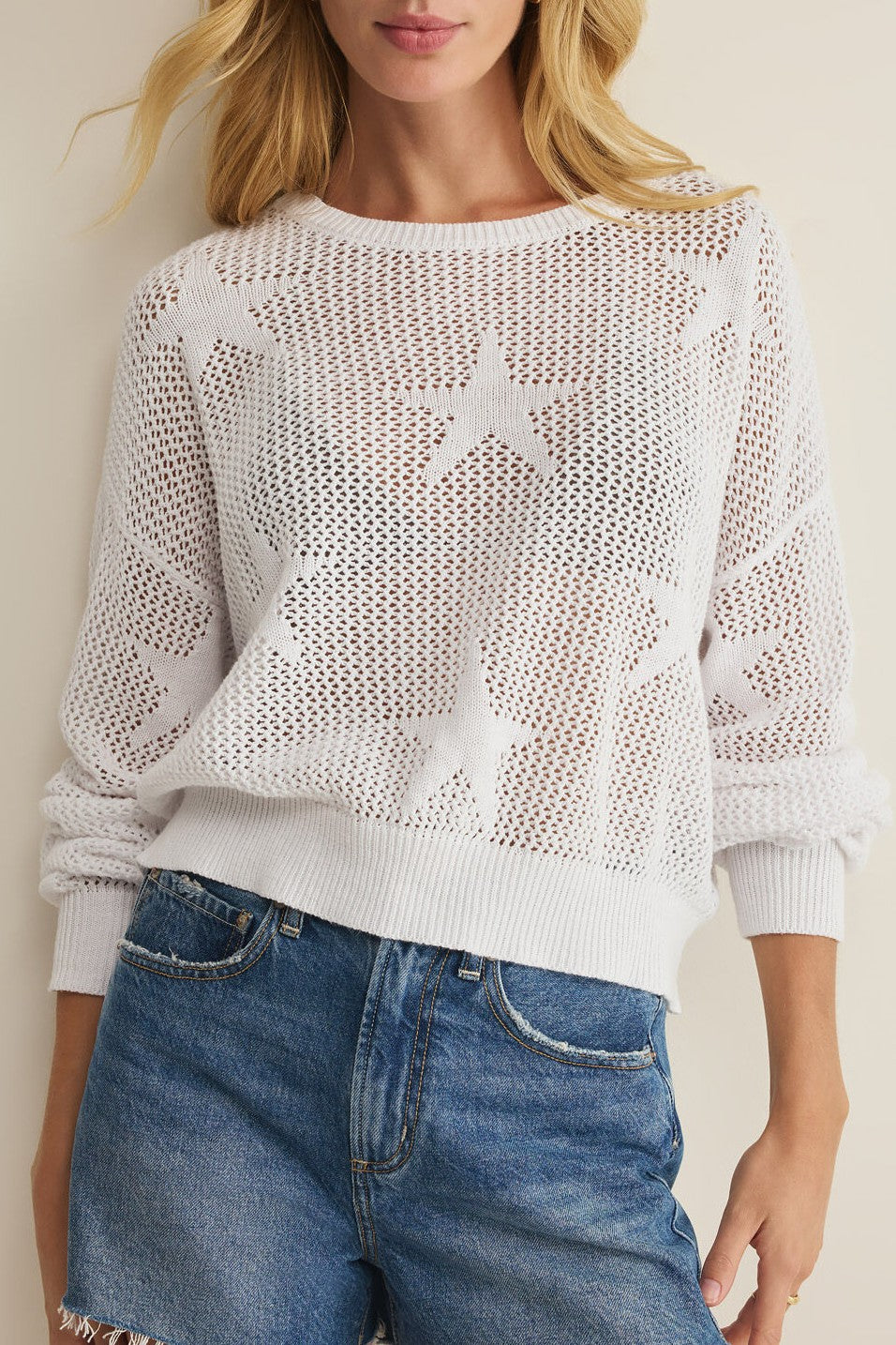 A woman wears the Z Supply "Star Of The Show Sweater" in white, a midweight long sleeve knit with star patterns, crew neck, ribbed hem and cuffs, over a white bra, paired with blue denim shorts.