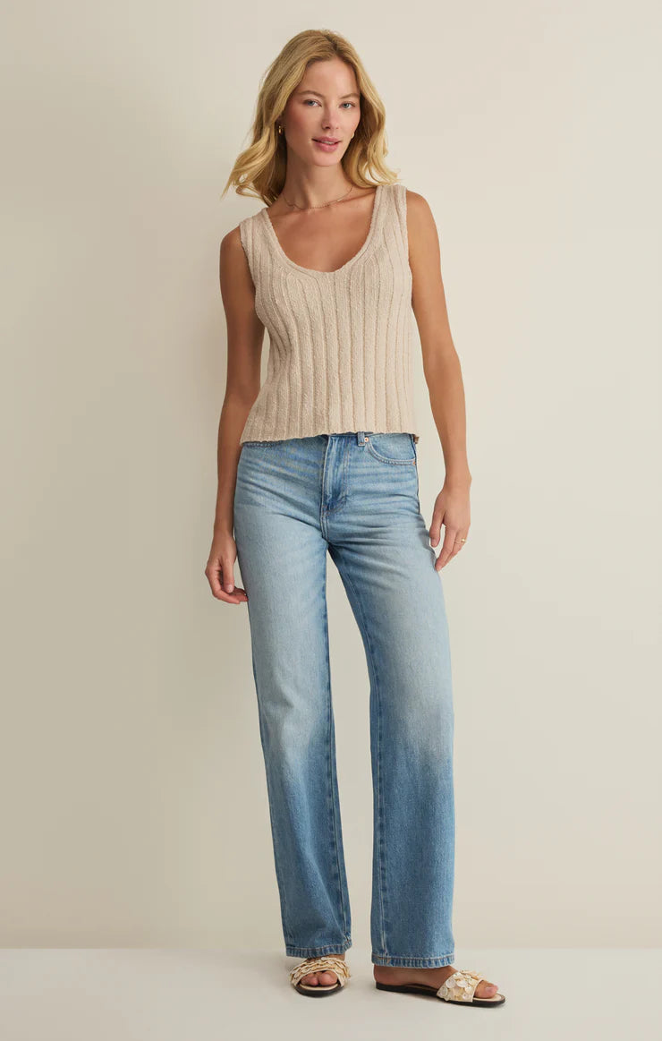 A woman with blonde hair poses casually against a plain background, wearing the Z Supply Acadia Sweater Tank in beige, light blue high-waisted jeans, and cream sandals.