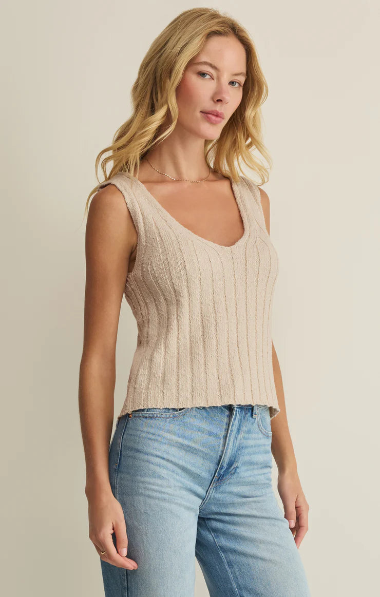 A woman with long blonde hair wears the Z Supply Acadia Sweater Tank, a sleeveless beige ribbed knit top with a V-neckline, paired with light blue jeans, standing against a plain light-colored background.