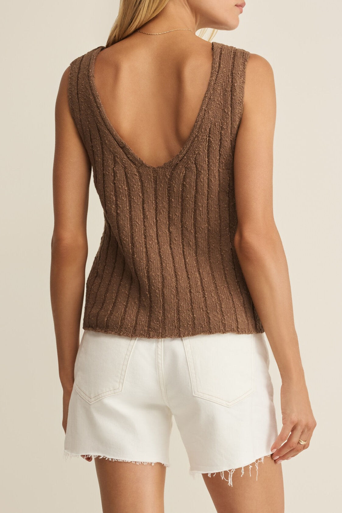 A woman stands against a neutral background wearing the Z Supply Acadia Sweater Tank, a sleeveless brown ribbed-knit V-neck with a low scoop back, paired with white frayed hem shorts.