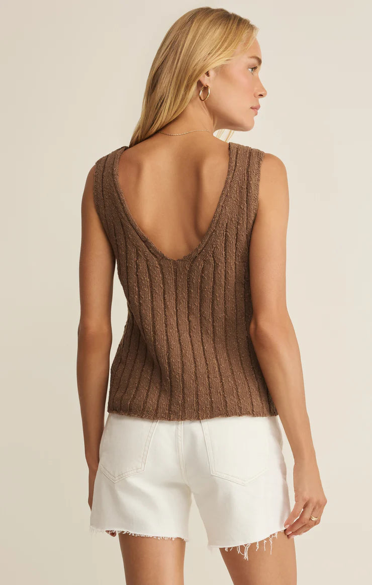 A woman with long blonde hair wears the Z Supply Acadia Sweater Tank, a sleeveless brown knit top with a deep scoop back, paired with white frayed-hem shorts. She stands facing away and looks to the side.