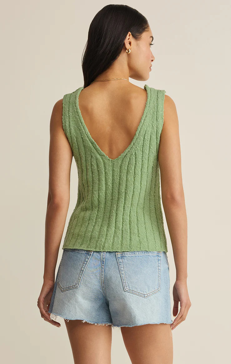 A woman faces away, wearing the Z Supply Acadia Sweater Tank—a sleeveless green knit top with a deep V-back—paired with light blue denim shorts against a neutral beige background.