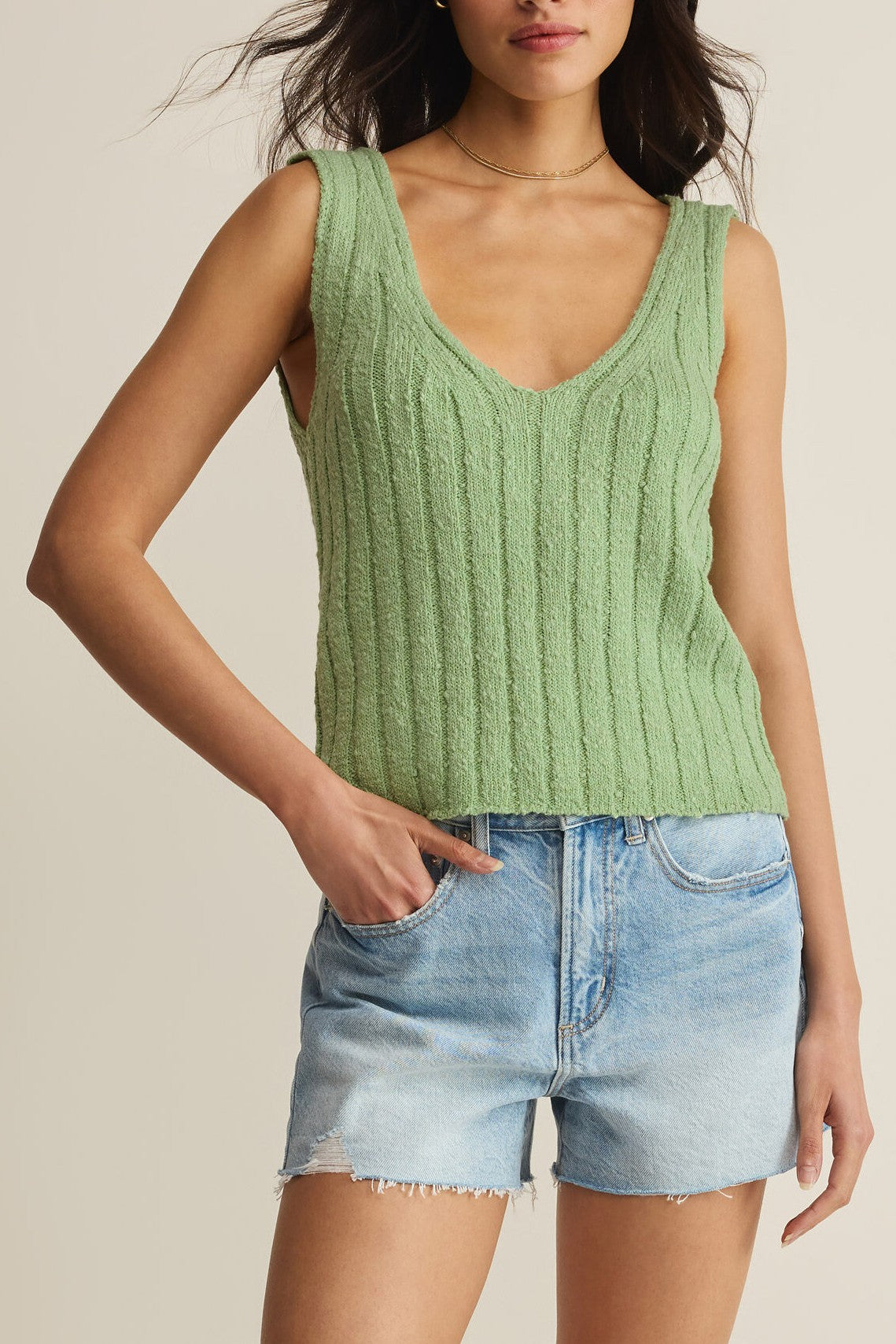 A woman wears the Z Supply Acadia Sweater Tank in green—sleeveless, ribbed, V-neck, and soft-washed—paired with light blue denim shorts with frayed hems, one hand in her pocket.