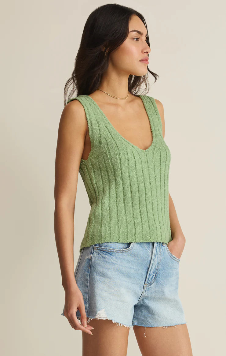 A woman with long dark hair stands with her hands in the pockets of light blue shorts, wearing the Z Supply Acadia Sweater Tank—a sleeveless, ribbed green top with a deep V-neck—looking to the side.