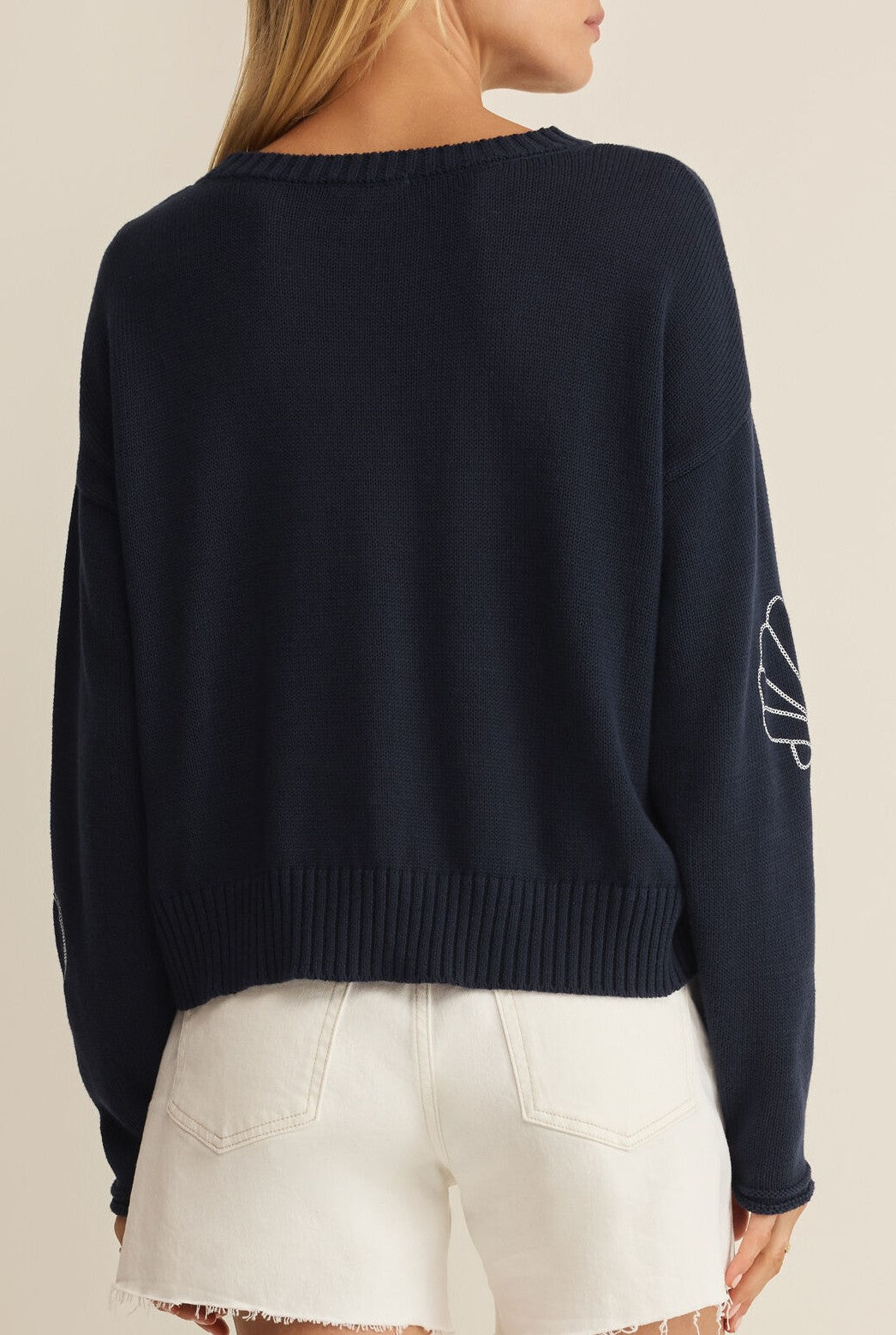 A woman faces away, wearing the Z Supply Paradise Reef Sienna Sweater—a lightweight cotton navy crew neck with ribbed hems and white elbow embroidery—paired with high-waisted frayed white denim shorts.