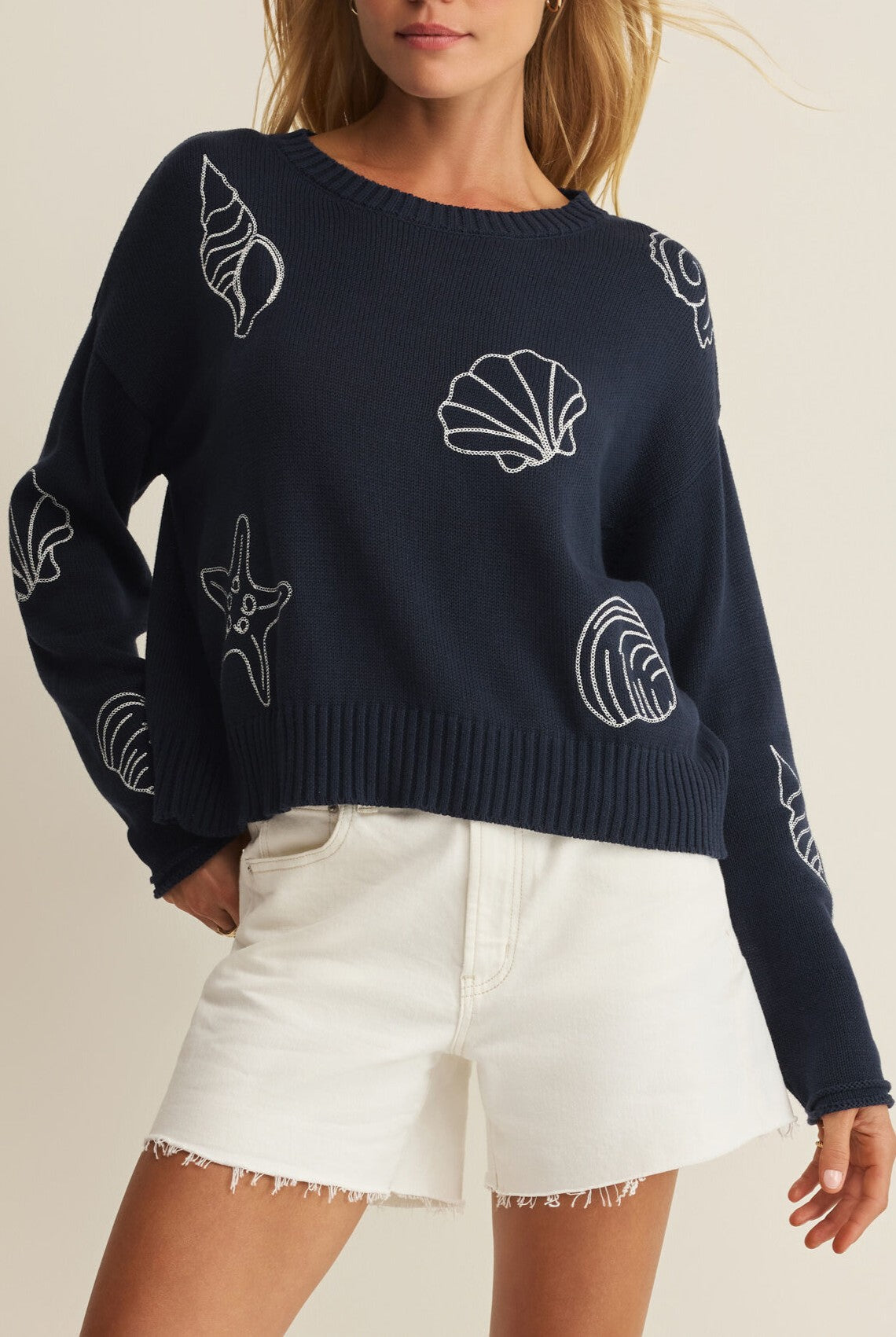 A woman wears the Z Supply Paradise Reef Sienna Sweater in navy featuring white seashell and starfish designs, styled with white high-waisted frayed denim shorts.