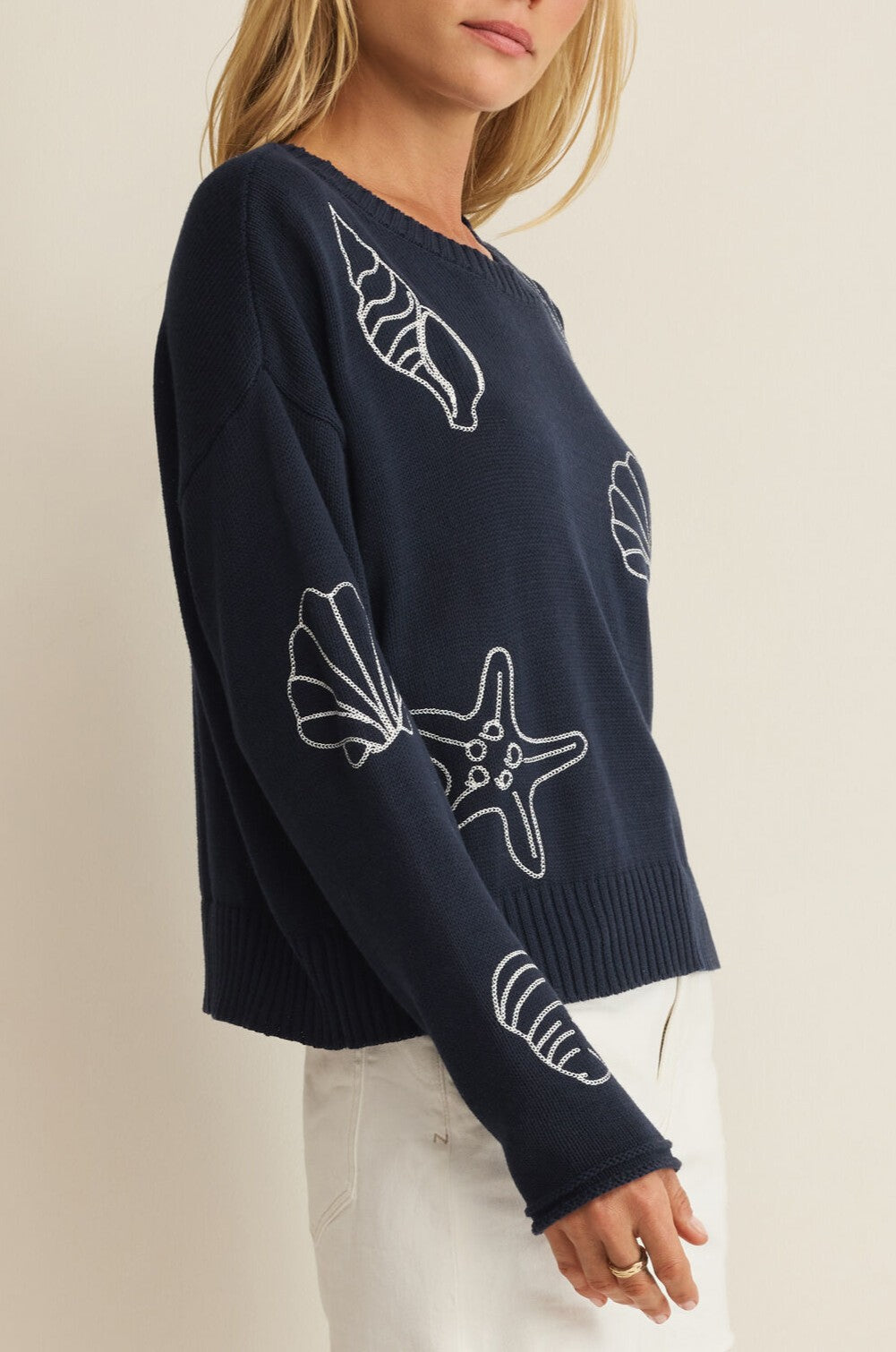 A woman wears the Z Supply Paradise Reef Sienna Sweater, a regular fit navy cotton top with white seashell and starfish designs, paired with white pants. She stands against a neutral background, her face partially out of view.