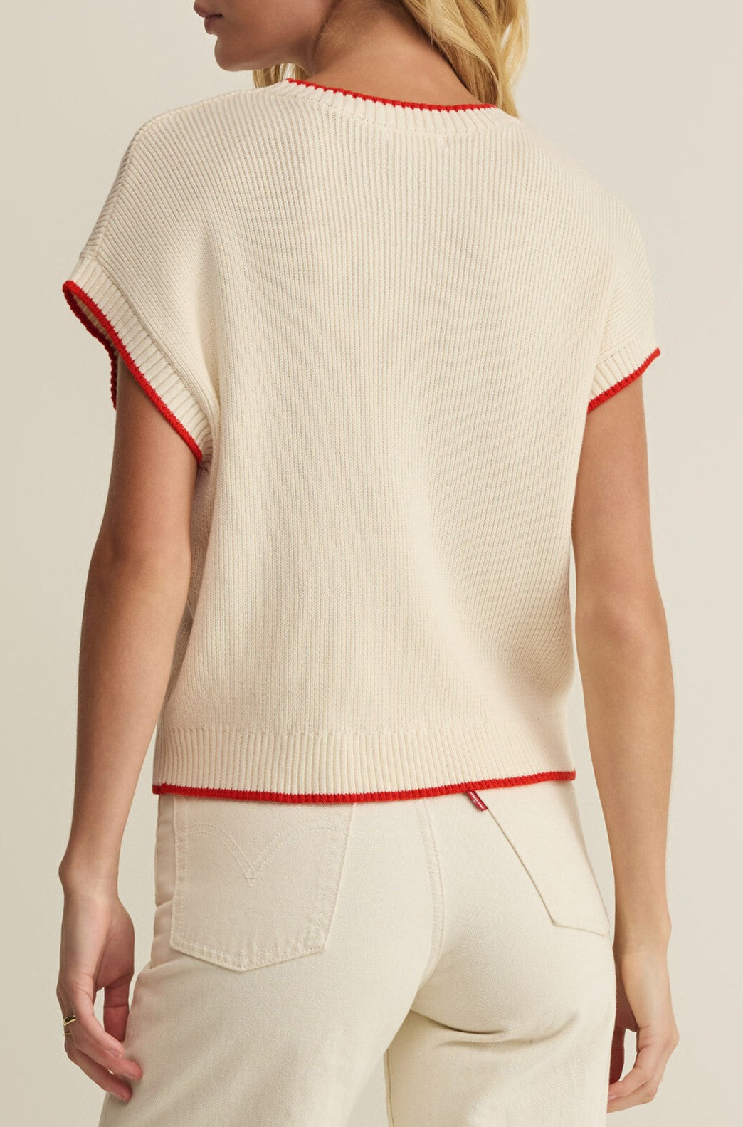 A woman is seen from the back wearing Z Supply's Sleeveless Patriot Sweater, a cream-colored knit top with red trim and ribbed detailing. Crafted from 100% cotton in a relaxed regular fit, she pairs it with high-waisted cream pants.