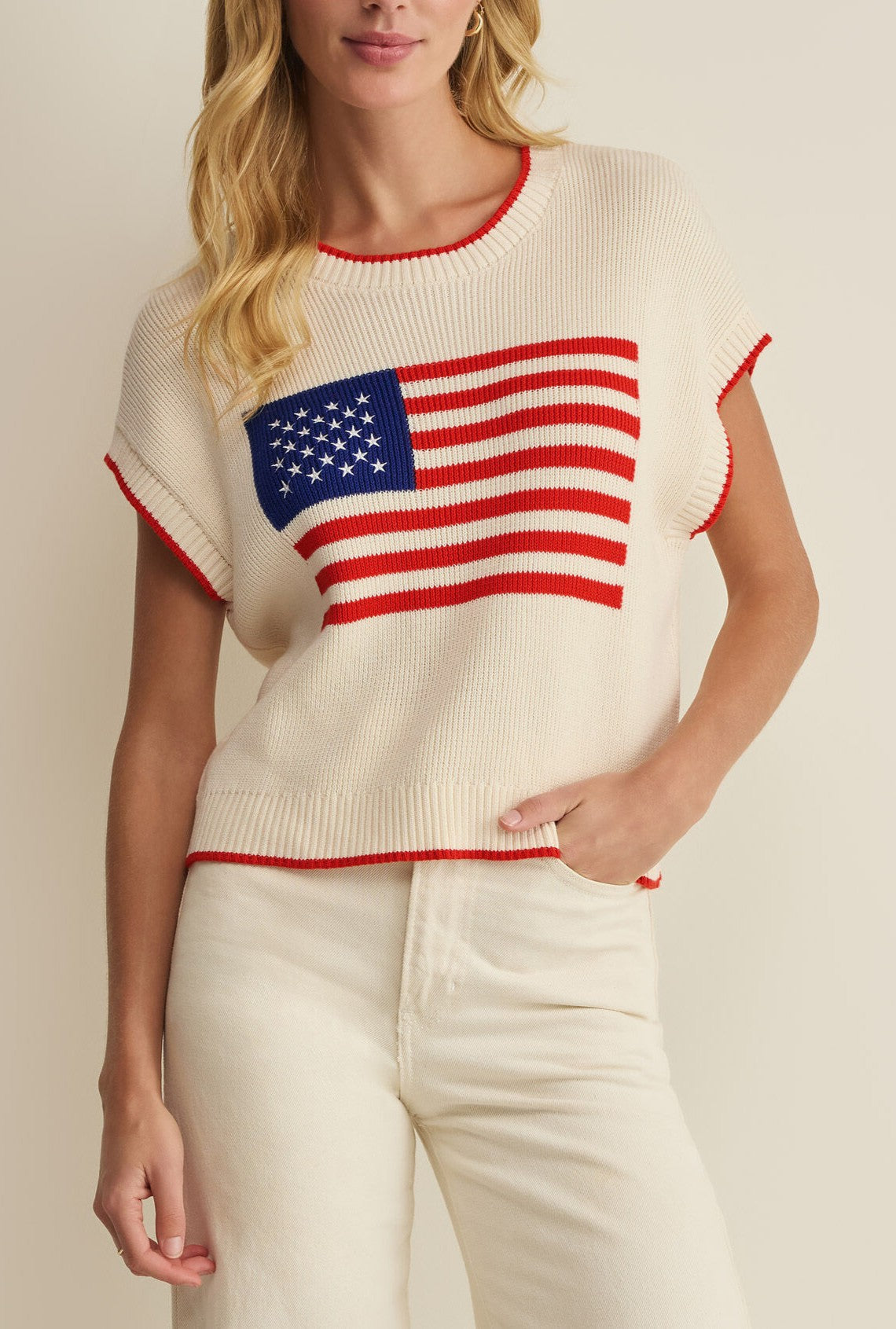 A woman models the Z Supply Sleeveless Patriot Sweater—a cream knit top with a bold American flag design—paired with cream pants, standing against a neutral background.
