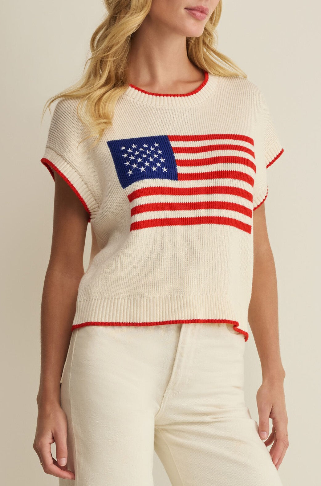A woman wearing the Z Supply Sleeveless Patriot Sweater, featuring an American flag design, paired with high-waisted cream pants.