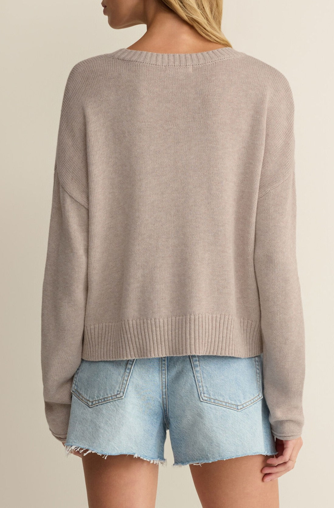 A person with light brown hair is seen from the back in a Z Supply Sienna Patriot Sweater—beige, 100% cotton, loose and long-sleeved—paired with light blue frayed-hem denim shorts against a neutral background.