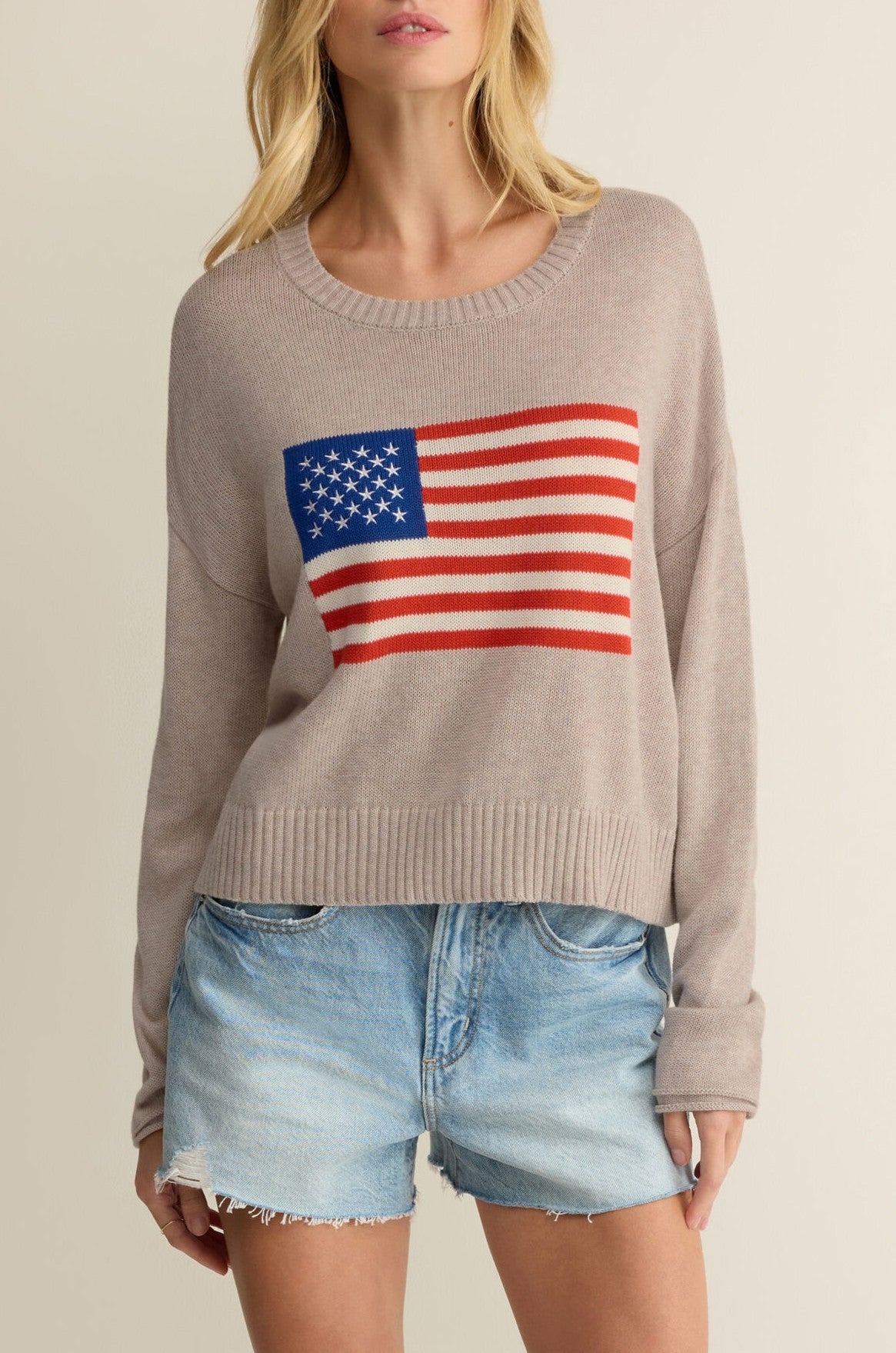 A woman wears the Z Supply Sienna Patriot Sweater in Tan Multi—a crew neck, long sleeve knit with an American flag graphic—paired with light blue denim shorts. The photo is cropped to show her lips and body only.