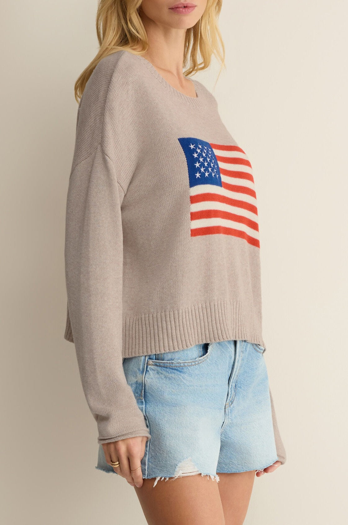 A woman in the Z Supply Sienna Patriot Sweater—100% cotton, crew neck, beige with an American flag on the front—and light blue denim shorts stands against a neutral background. Her face is partially out of frame.