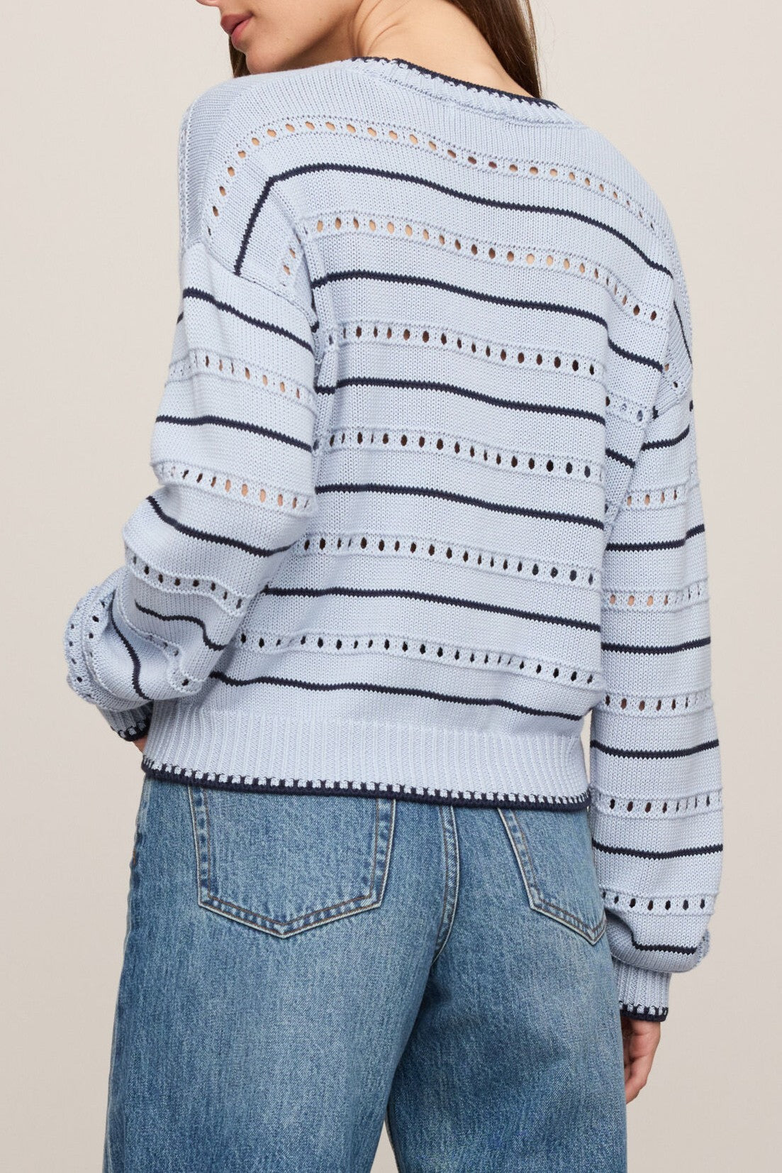 A woman is seen from behind wearing the Z Supply Darby Stripe Sweater—a light blue, regular fit crew neck knit with horizontal navy stripes and eyelet details—paired with high-waisted blue jeans. The sweater features long sleeves, and the photo highlights the back of her outfit.