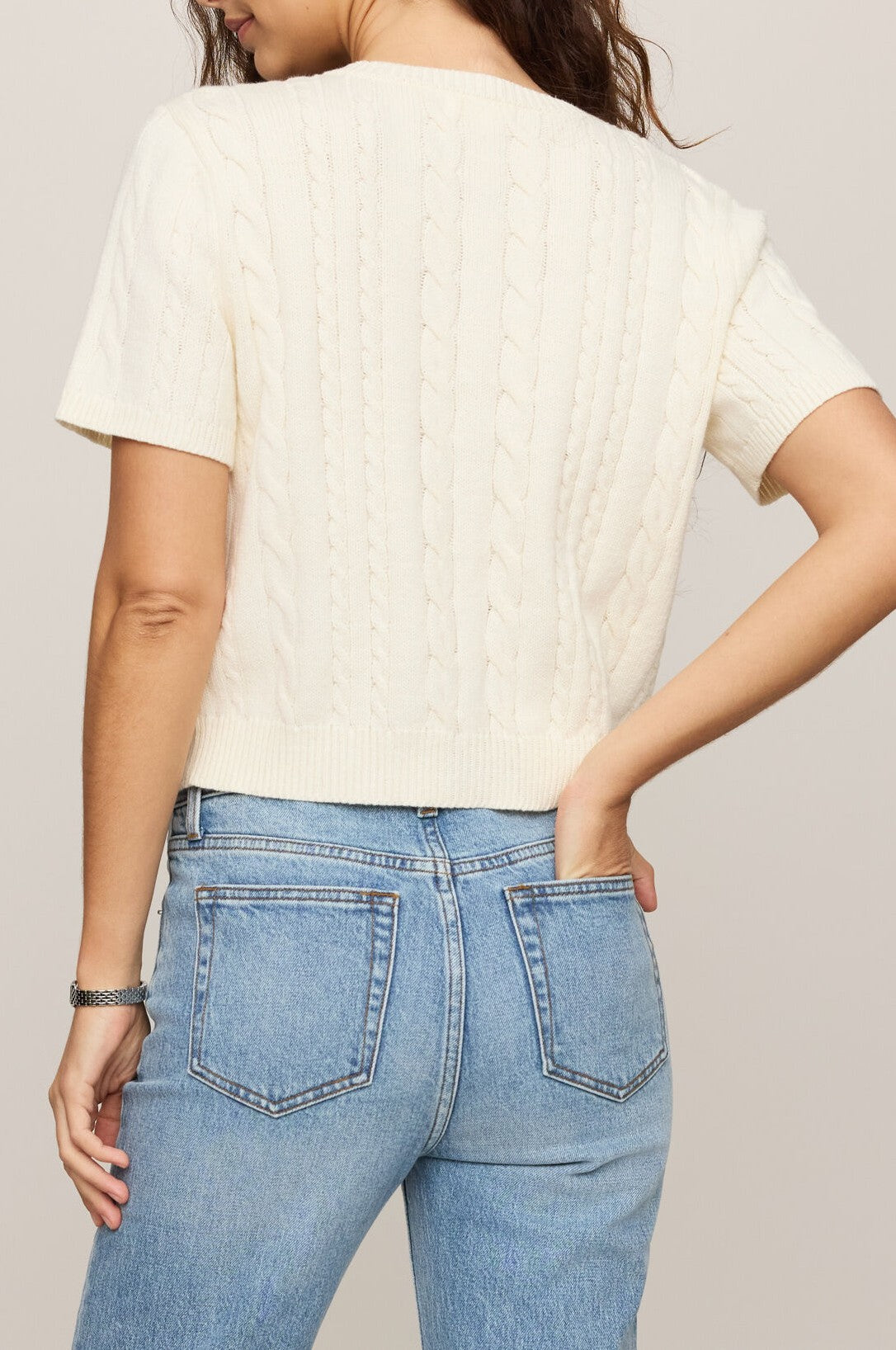 A woman is shown from behind wearing the Z Supply Cera Cardigan, a cream-colored, short-sleeved cable knit sweater with a crew neck, paired with high-waisted blue jeans and one hand resting on her hip.