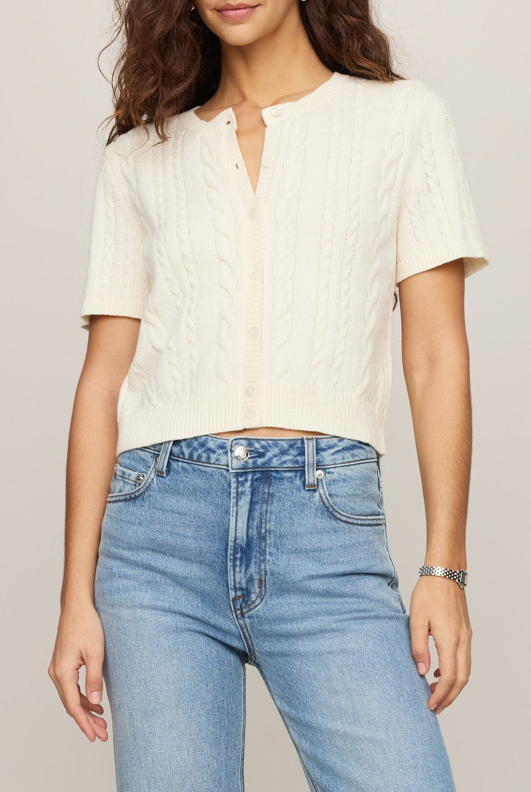 A woman wearing the Z Supply Cera Cardigan in CLOUD—a short-sleeve, cable-knit button front cardigan with a crew neckline—paired with high-waisted blue jeans. She has wavy brown hair and a silver bracelet on her wrist. The photo is cropped at the shoulders and thighs.
