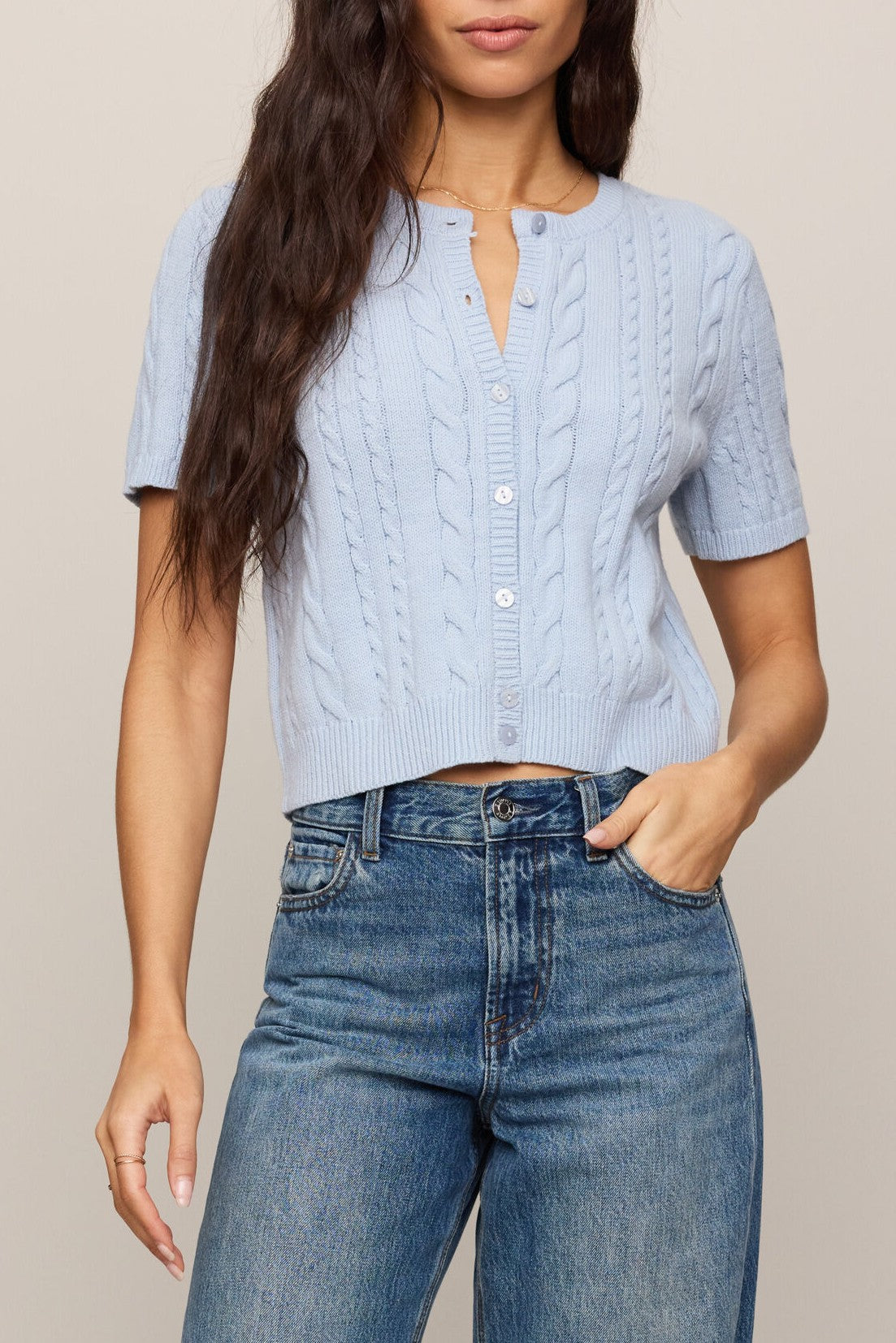 A woman wears the Z Supply Cera Cardigan in SKYLINE, a light blue regular fit short-sleeve cable-knit button-up sweater, paired with high-waisted blue jeans. Her hand rests in her pocket, and her long brown hair is loose.