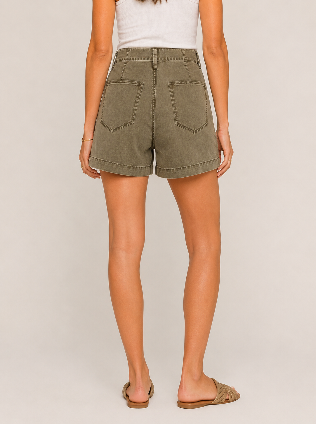 A woman faces away, wearing Heartloom's Perri Twill Short in olive green with a high waist, zipper closure, and two large back pockets, paired with a white tank top and tan slide sandals against a plain light background.