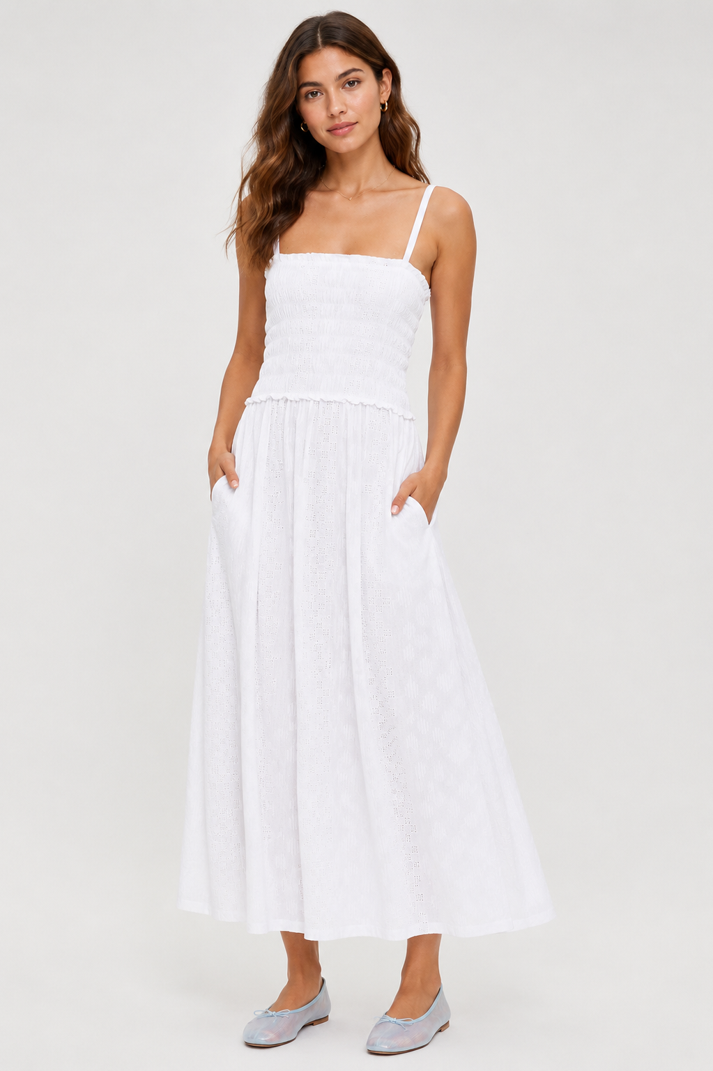 A woman with long brown hair stands against a light background, wearing the Splendid Kendra Geo Eyelet Dress—an ankle-length, sleeveless white dress with a smocked bodice and flowing skirt—paired with flat light-colored shoes.