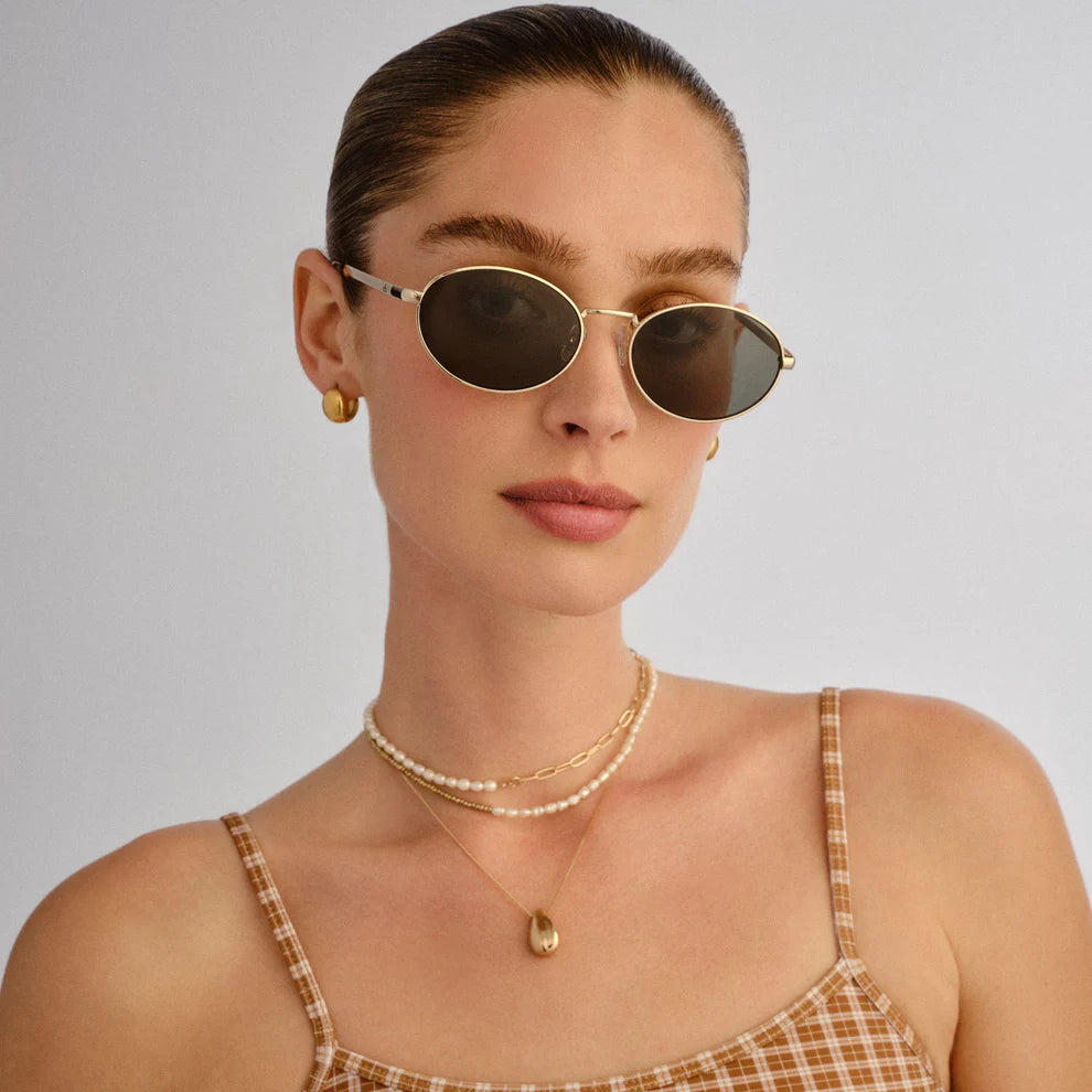 A woman with slicked-back hair models Aire's Ephermeris Sunnies—round gold frame sunglasses—paired with gold earrings, layered necklaces, and a brown plaid spaghetti-strap top against a light background.