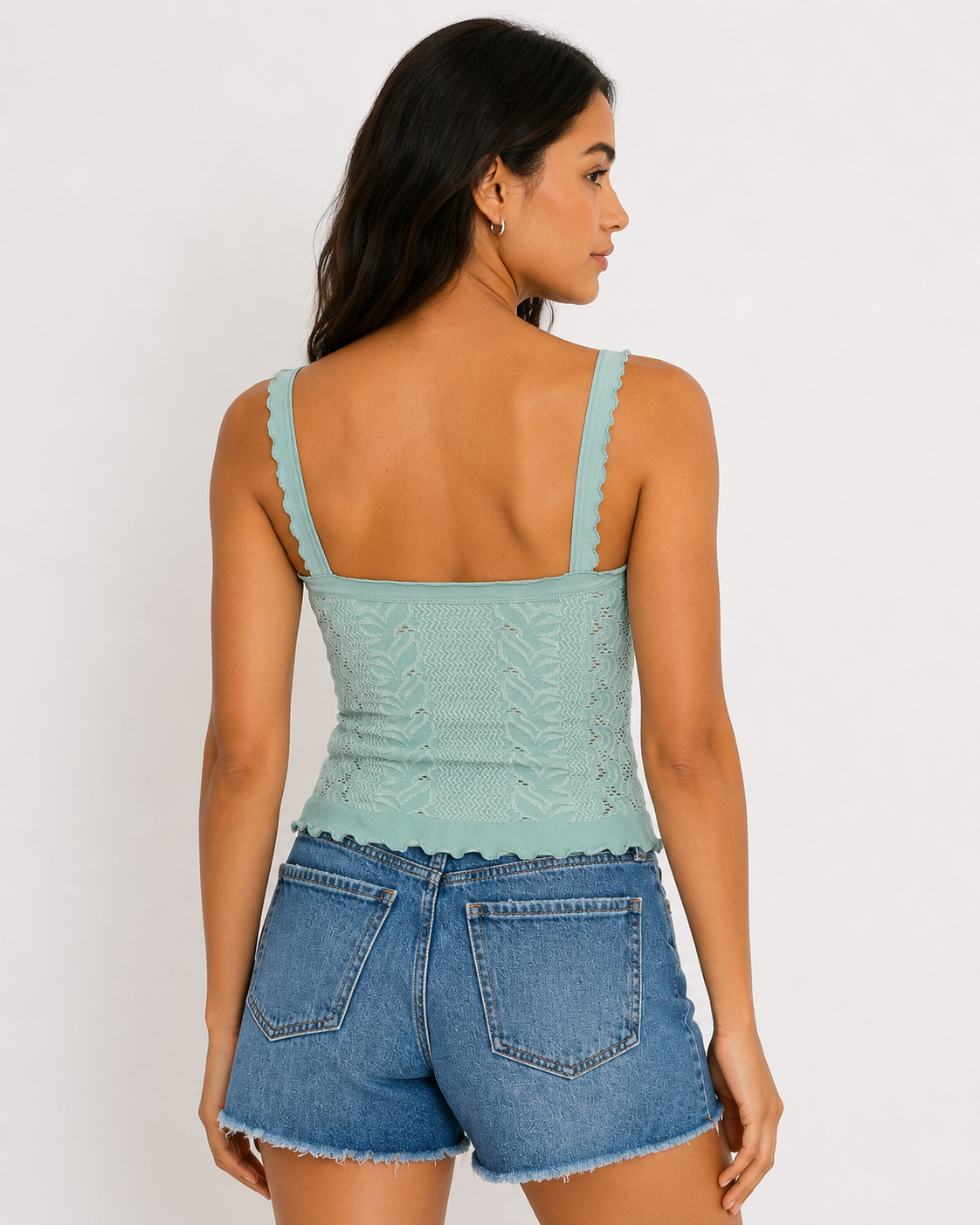 A woman with long dark hair faces away, wearing the Free People So Soft Femme Square Top featuring lace and lettuce edge details, paired with high-waisted frayed hem denim shorts. The background is plain and light-colored.