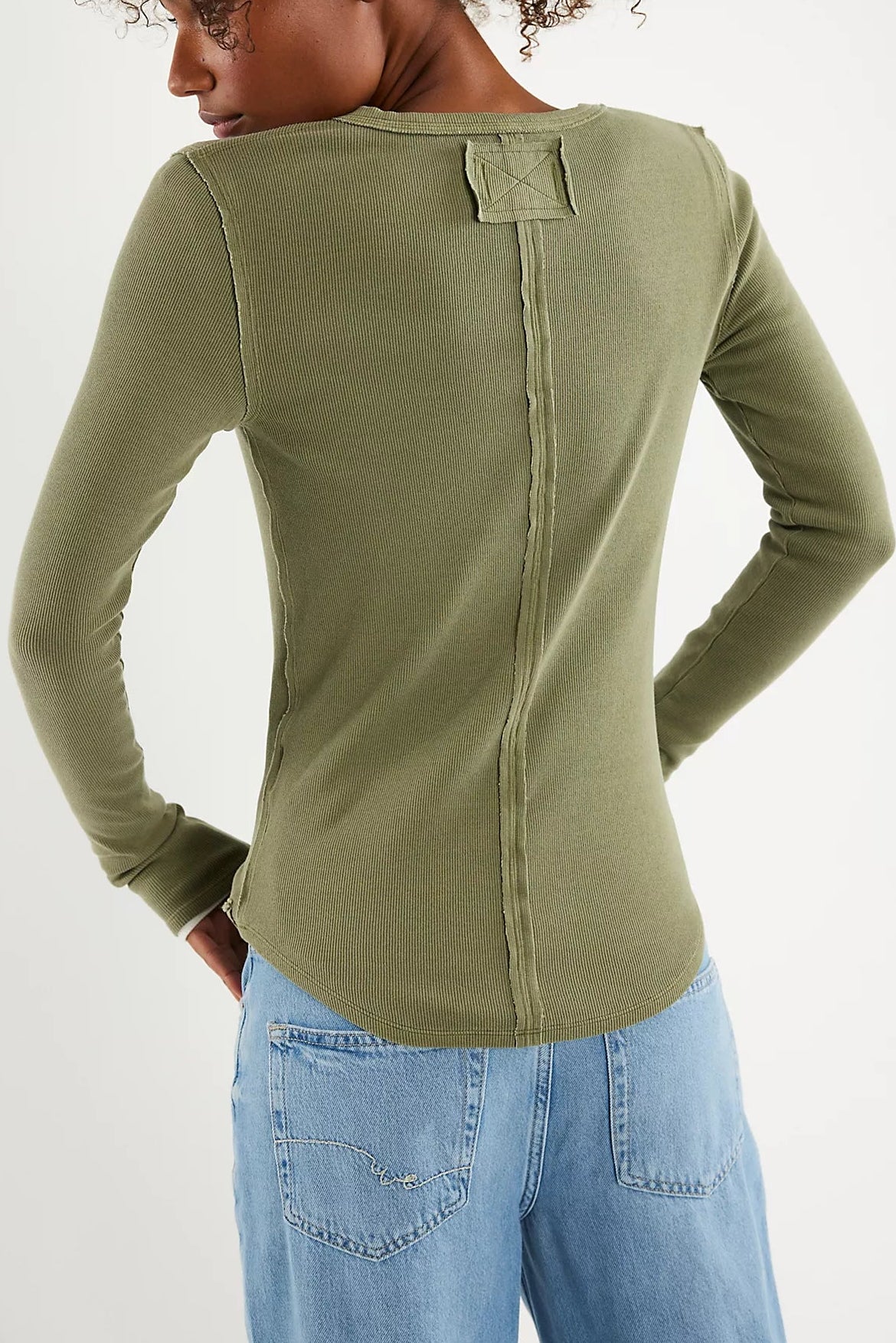 A person wearing the Free People Jamie Henley in Capulet Olive—a fitted olive green cotton blend shirt with exposed seams and crewneck long sleeves—paired with light blue jeans, shown from the back with hands on hips.