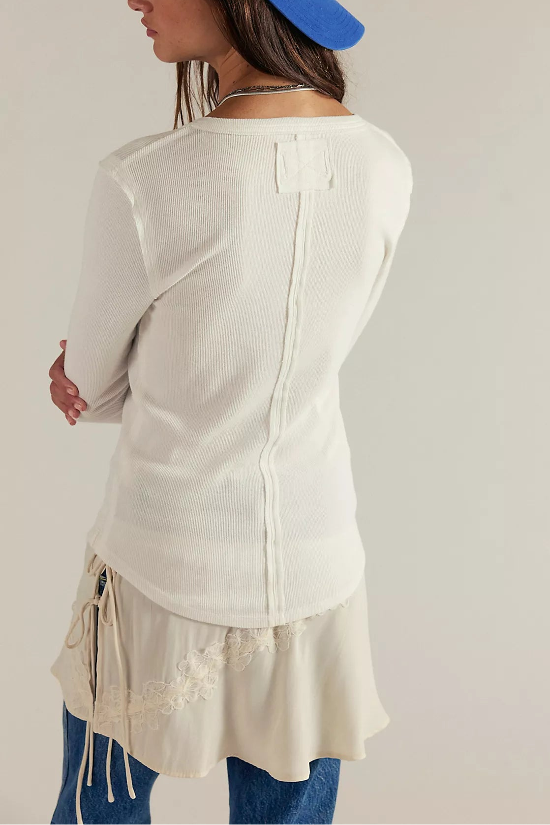 A person wearing the Free People Jamie Henley In Ivory—a long sleeve, crewneck cotton top with a button half placket—paired with a cream embroidered skirt, blue jeans, and a blue hat, is shown from the back with arms crossed.