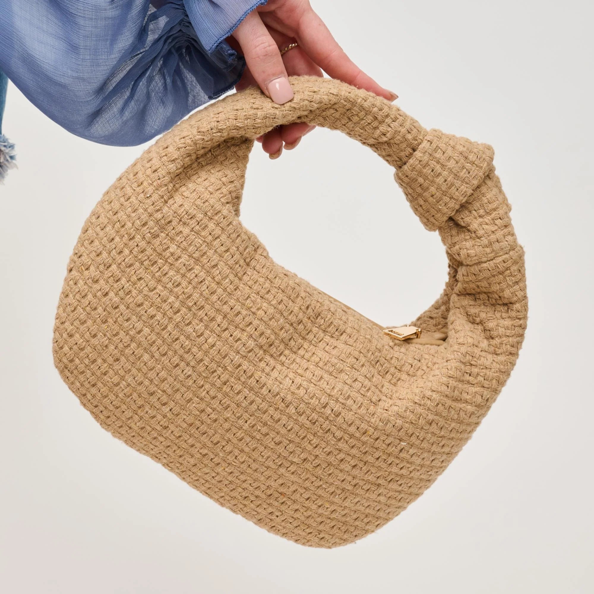 A hand holds the Urban Expressions Tracy Bag in Natural Tweed, a chic tan handbag with a knotted, textured handle, against a plain light background. The person is wearing a blue long-sleeved top.