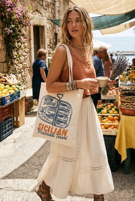 A woman in a sleeveless top and long skirt strolls through an outdoor market, carrying a South Moon Under Canvas Tote and a bundle of lavender, with fruit stands and people in the background.