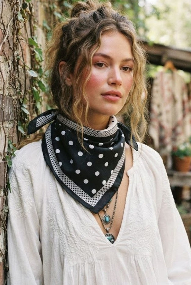A young woman with wavy hair wears a white blouse and delicately layered necklaces, accessorized with the South Moon Under Polka Dot Scarf in BLACK around her neck. She stands outdoors near a wall draped in climbing plants, with blurred greenery in the background.