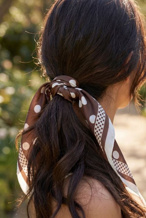 A woman with long, wavy brown hair styled in a low ponytail, secured with the South Moon Under Polka Dot Scarf in BROWN, enjoying the natural sunlight outdoors.